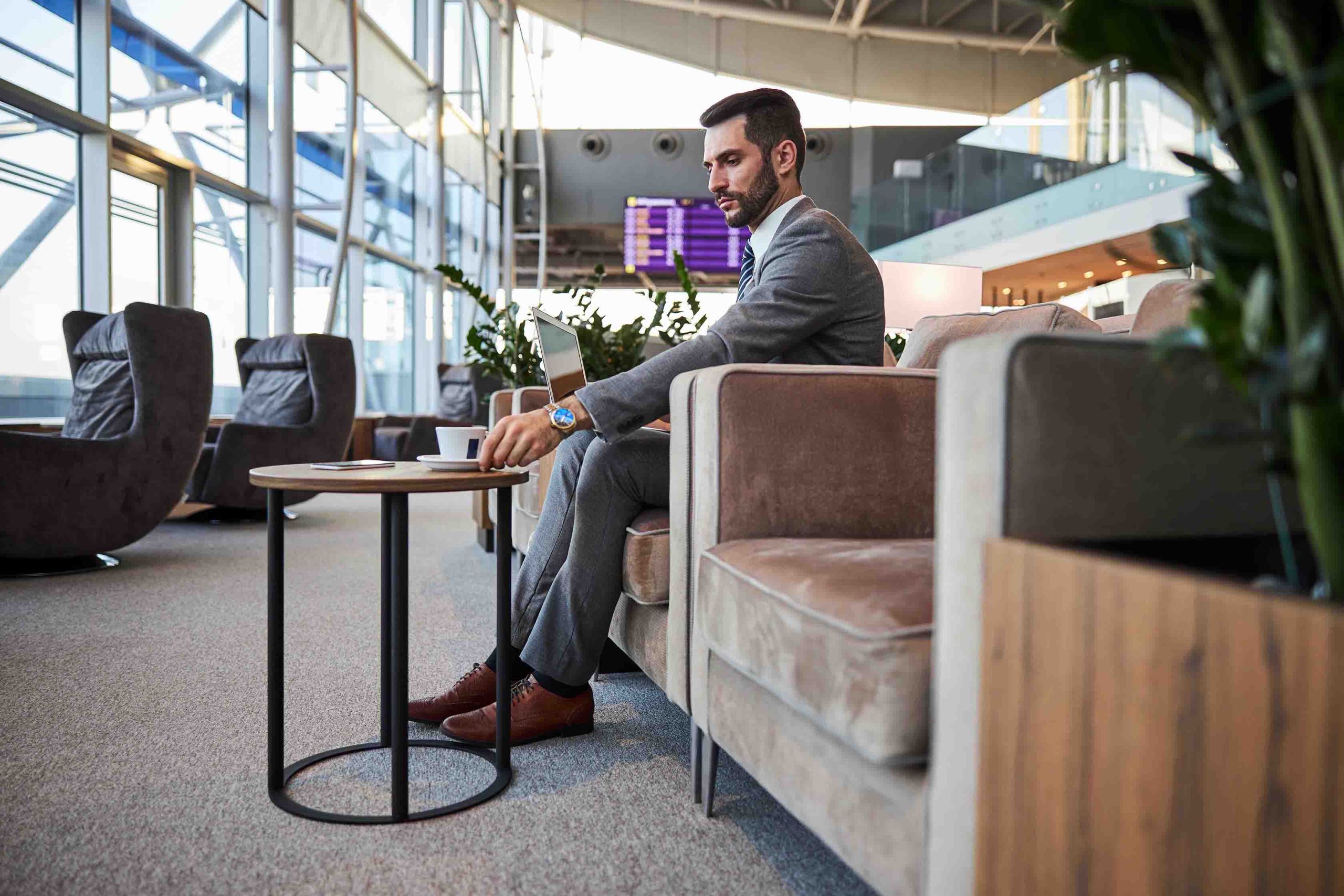 Handsome businessman with a laptop sitting in the armchair in lounge zone and putting a cup on a coffee table